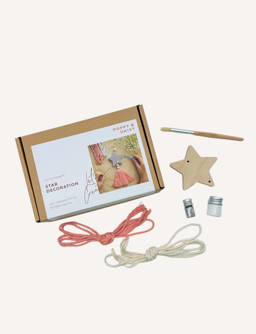 A child is busy with a Star Decoration craft project by Poppy & Daisy, featuring a glittery star, pink tassel, and wooden beads. Nearby lie pink and white cords, white flowers, and a needle on a round wooden surface.