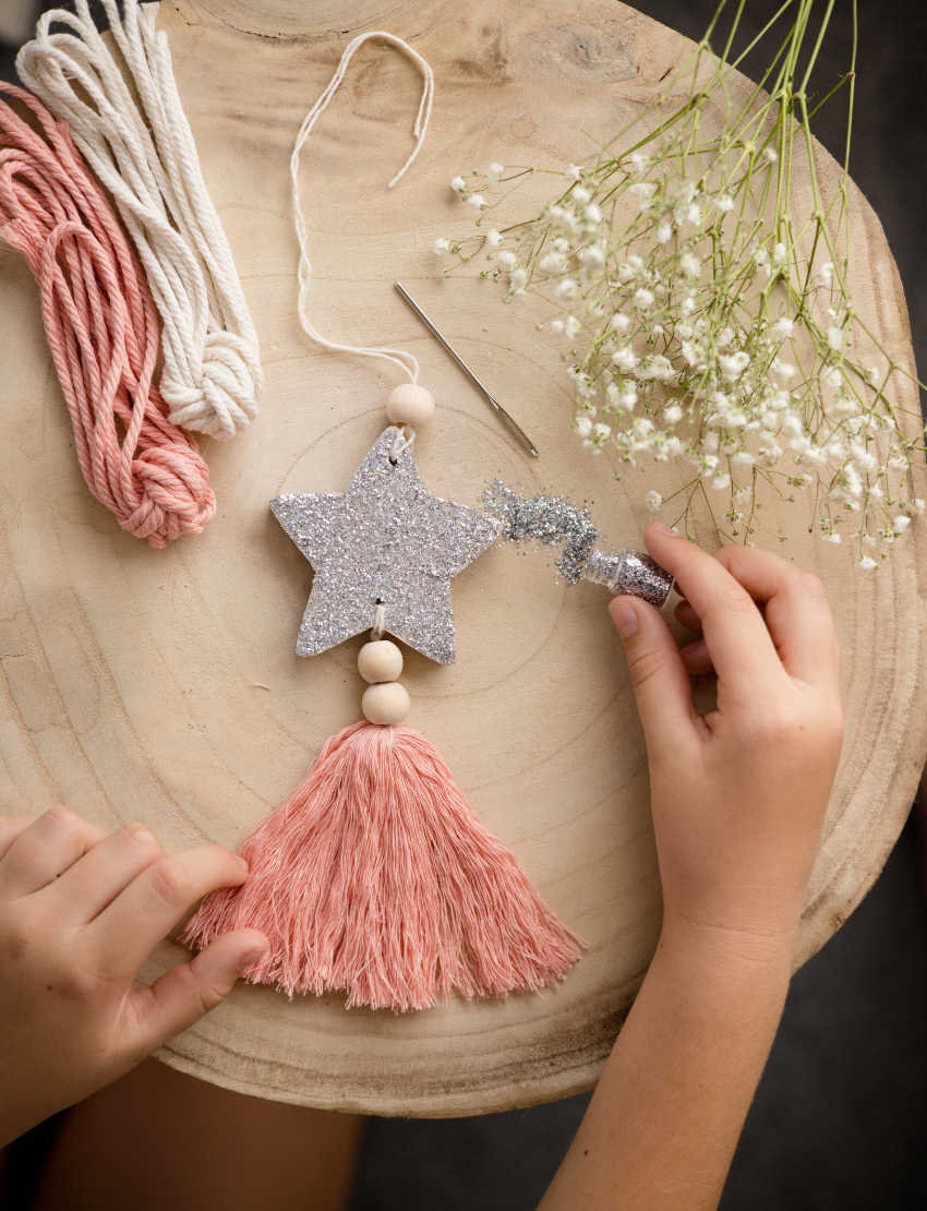 A child is busy with a Star Decoration craft project by Poppy & Daisy, featuring a glittery star, pink tassel, and wooden beads. Nearby lie pink and white cords, white flowers, and a needle on a round wooden surface.