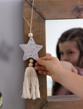 A child is busy with a Star Decoration craft project by Poppy & Daisy, featuring a glittery star, pink tassel, and wooden beads. Nearby lie pink and white cords, white flowers, and a needle on a round wooden surface.