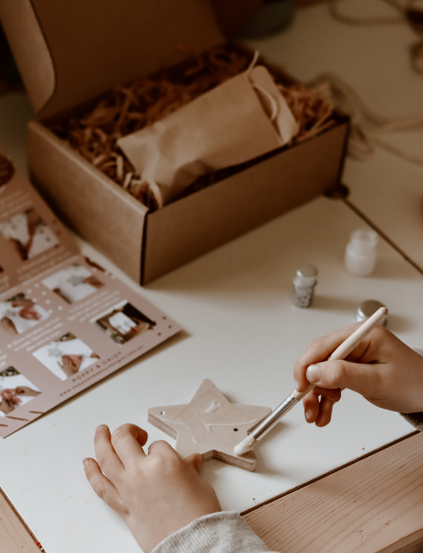 A child is busy with a Star Decoration craft project by Poppy & Daisy, featuring a glittery star, pink tassel, and wooden beads. Nearby lie pink and white cords, white flowers, and a needle on a round wooden surface.