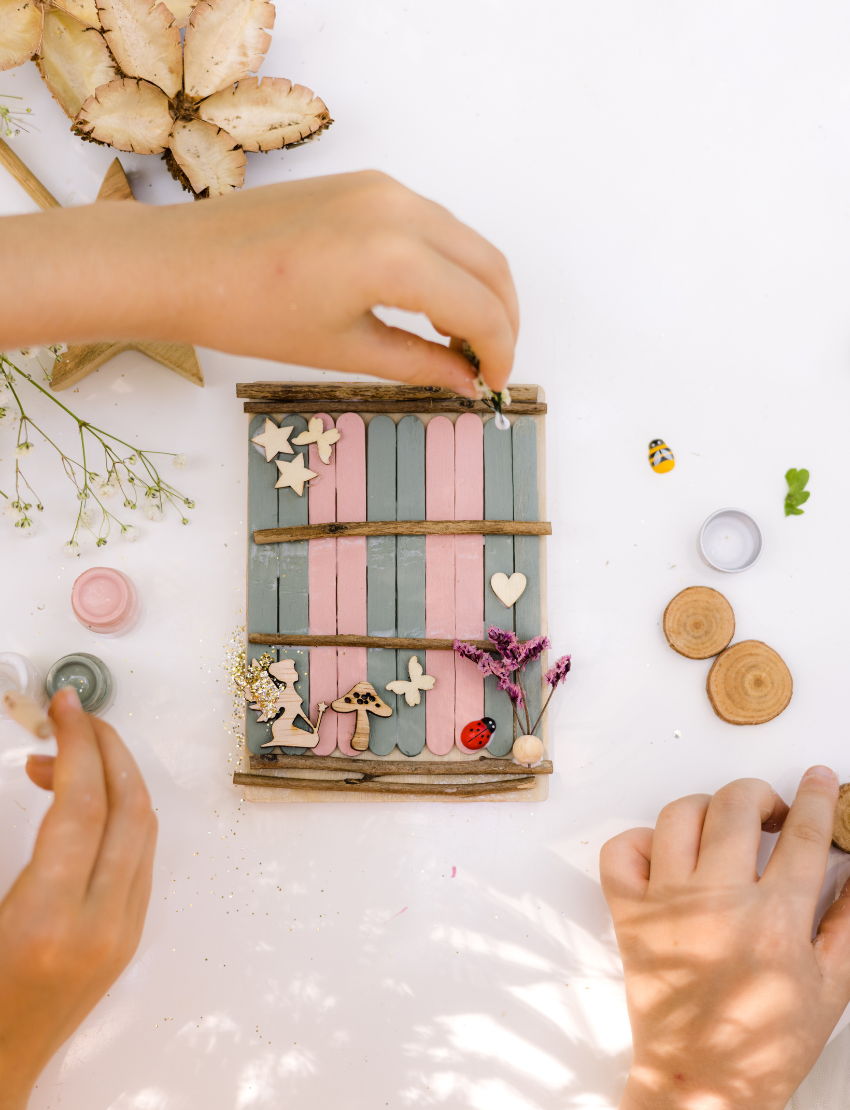 Hands decorate a delicate Fairy Door by Poppy & Daisy, crafted from a small, colorful fence of blue and pink popsicle sticks adorned with wooden stars, flowers, and other embellishments. Nearby are paint jars, brushes, and wooden disks on a white surface sprinkled with dried flowers.