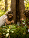 Hands decorate a delicate Fairy Door by Poppy & Daisy, crafted from a small, colorful fence of blue and pink popsicle sticks adorned with wooden stars, flowers, and other embellishments. Nearby are paint jars, brushes, and wooden disks on a white surface sprinkled with dried flowers.