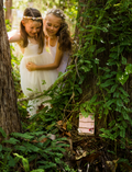 Hands decorate a delicate Fairy Door by Poppy & Daisy, crafted from a small, colorful fence of blue and pink popsicle sticks adorned with wooden stars, flowers, and other embellishments. Nearby are paint jars, brushes, and wooden disks on a white surface sprinkled with dried flowers.