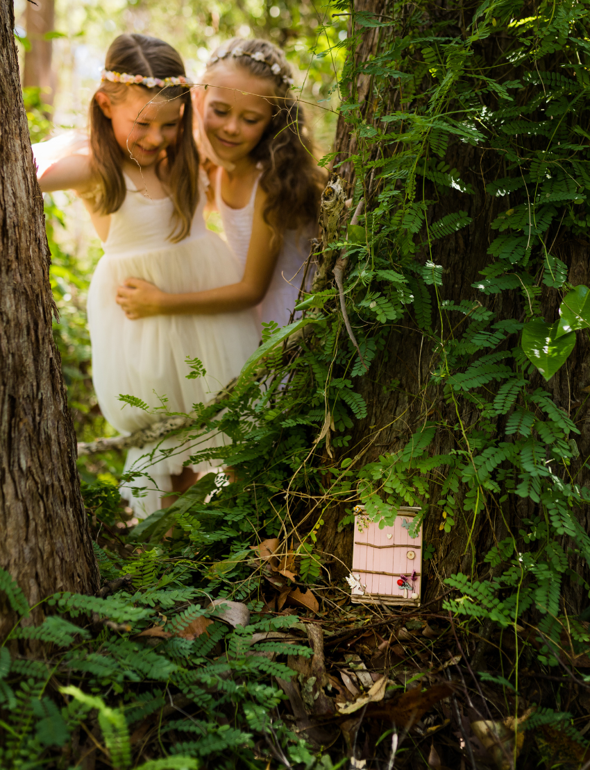 Hands decorate a delicate Fairy Door by Poppy & Daisy, crafted from a small, colorful fence of blue and pink popsicle sticks adorned with wooden stars, flowers, and other embellishments. Nearby are paint jars, brushes, and wooden disks on a white surface sprinkled with dried flowers.