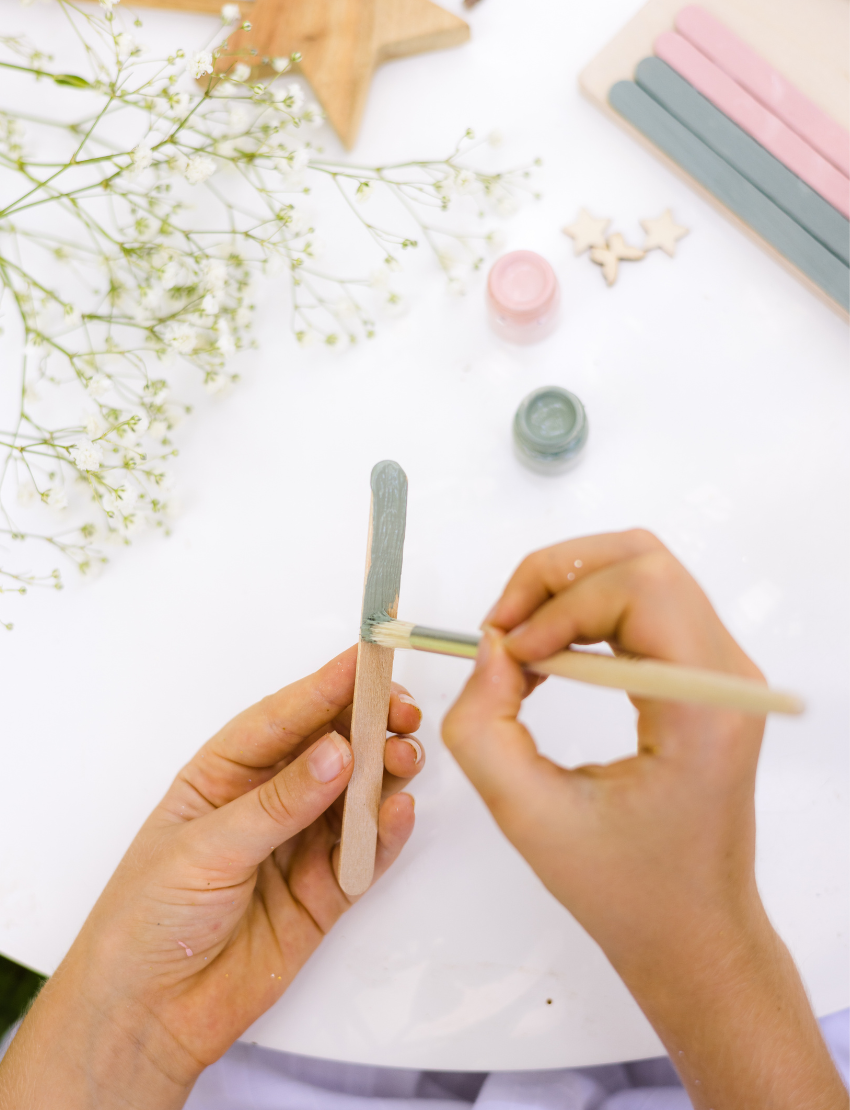 Hands decorate a delicate Fairy Door by Poppy & Daisy, crafted from a small, colorful fence of blue and pink popsicle sticks adorned with wooden stars, flowers, and other embellishments. Nearby are paint jars, brushes, and wooden disks on a white surface sprinkled with dried flowers.