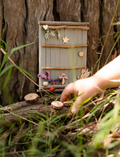 Hands decorate a delicate Fairy Door by Poppy & Daisy, crafted from a small, colorful fence of blue and pink popsicle sticks adorned with wooden stars, flowers, and other embellishments. Nearby are paint jars, brushes, and wooden disks on a white surface sprinkled with dried flowers.