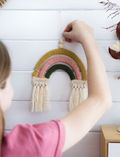 A person in a pink shirt hangs the Poppy & Daisy Macrame Rainbow, crafted with yarn and tassels, on a white wooden wall. The decoration showcases mustard yellow, pink, and green colors.