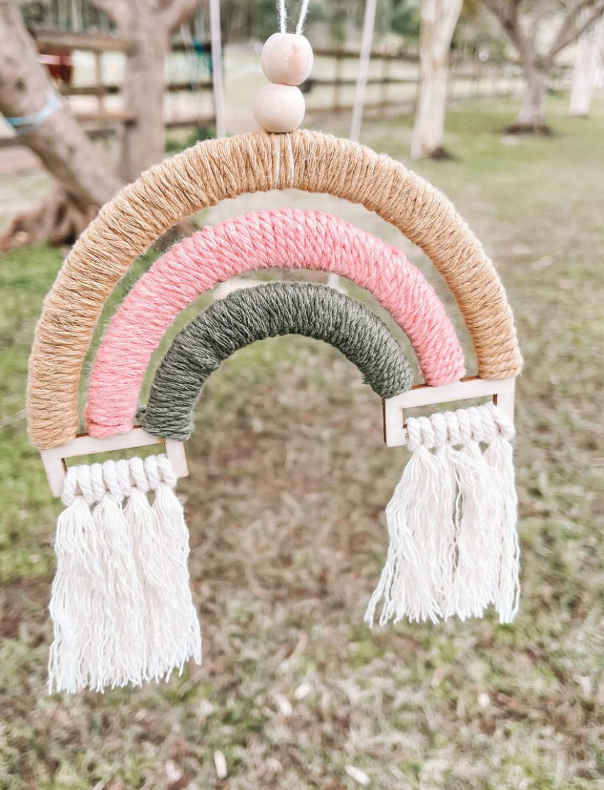 A person in a pink shirt hangs the Poppy & Daisy Macrame Rainbow, crafted with yarn and tassels, on a white wooden wall. The decoration showcases mustard yellow, pink, and green colors.