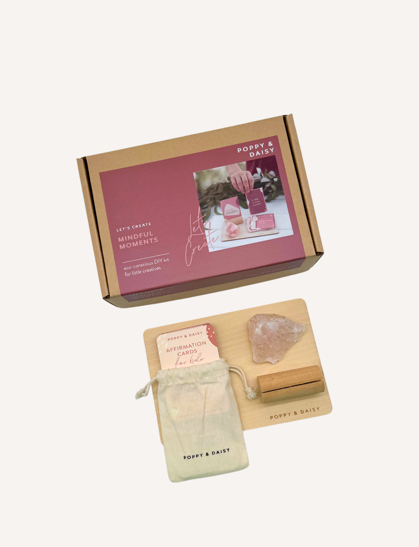 A person holds a set of Mindful Moments cards by Poppy & Daisy, featuring abstract designs and text. Nearby, a wooden tray contains a pink crystal, a small box, and additional cards. A cardboard box displaying the Poppy & Daisy branding rests on a textured surface.