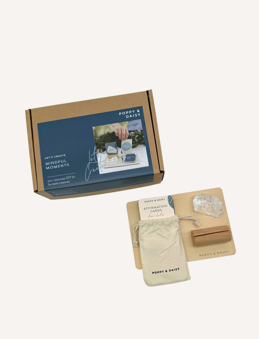 A person holds a set of Mindful Moments cards by Poppy & Daisy, featuring abstract designs and text. Nearby, a wooden tray contains a pink crystal, a small box, and additional cards. A cardboard box displaying the Poppy & Daisy branding rests on a textured surface.