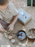 In an outdoor setting filled with natural light, a child is immersed in play on a sandy surface with Poppy & Daisy's Fun Fossils set, featuring wooden bowls and utensils. Nearby lies an open cardboard box adorned with the product image on its lid.