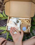 In an outdoor setting filled with natural light, a child is immersed in play on a sandy surface with Poppy & Daisy's Fun Fossils set, featuring wooden bowls and utensils. Nearby lies an open cardboard box adorned with the product image on its lid.