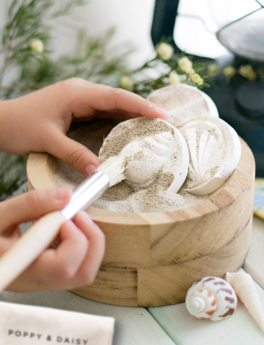 In an outdoor setting filled with natural light, a child is immersed in play on a sandy surface with Poppy & Daisy's Fun Fossils set, featuring wooden bowls and utensils. Nearby lies an open cardboard box adorned with the product image on its lid.