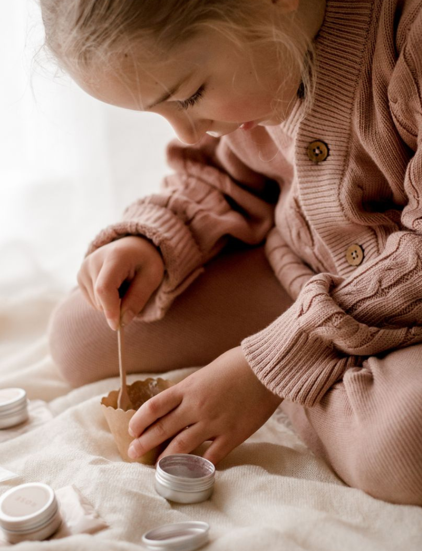 A young girl with long hair holds a Poppy & Daisy Mineral Makeup brush and gazes into a mirror. Kneeling on the floor, she wears a pink sweater and leggings, while various small containers of Mineral Makeup by Poppy & Daisy are arranged on a nearby white stool. The setting is bright and airy.