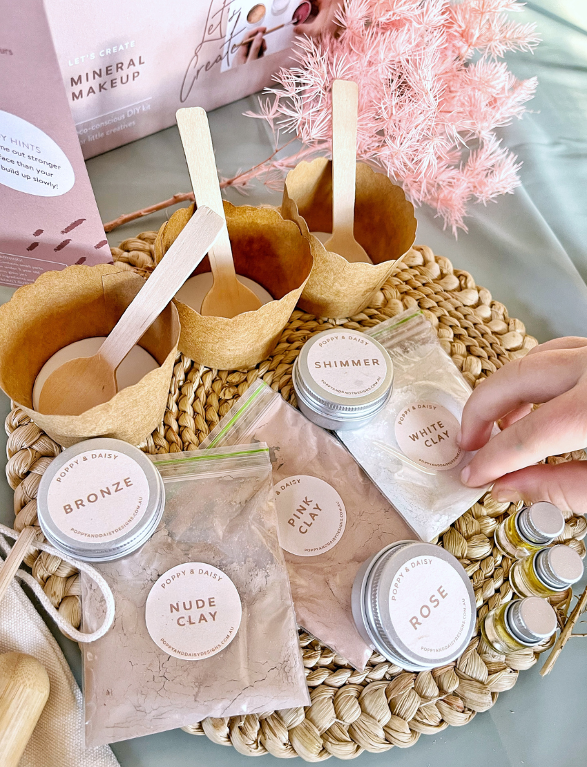 A young girl with long hair holds a Poppy & Daisy Mineral Makeup brush and gazes into a mirror. Kneeling on the floor, she wears a pink sweater and leggings, while various small containers of Mineral Makeup by Poppy & Daisy are arranged on a nearby white stool. The setting is bright and airy.