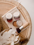 A young girl with long hair holds a Poppy & Daisy Mineral Makeup brush and gazes into a mirror. Kneeling on the floor, she wears a pink sweater and leggings, while various small containers of Mineral Makeup by Poppy & Daisy are arranged on a nearby white stool. The setting is bright and airy.