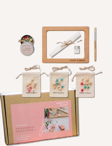 Hands are threading wooden beads onto cotton cords over a frame labeled "Poppy & Daisy," surrounded by small cloth bags filled with beads and a pink bougainvillea flower on a white surface, creating a charming scene for the Poppy & Daisy Friendship Necklaces kit.