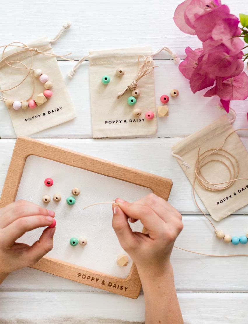 Hands are threading wooden beads onto cotton cords over a frame labeled "Poppy & Daisy," surrounded by small cloth bags filled with beads and a pink bougainvillea flower on a white surface, creating a charming scene for the Poppy & Daisy Friendship Necklaces kit.