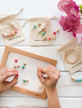 Hands are threading wooden beads onto cotton cords over a frame labeled 