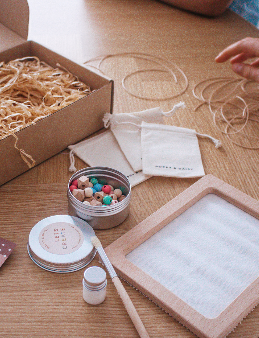 Hands are threading wooden beads onto cotton cords over a frame labeled "Poppy & Daisy," surrounded by small cloth bags filled with beads and a pink bougainvillea flower on a white surface, creating a charming scene for the Poppy & Daisy Friendship Necklaces kit.