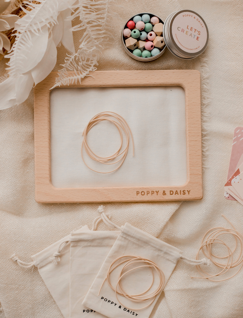 Hands are threading wooden beads onto cotton cords over a frame labeled "Poppy & Daisy," surrounded by small cloth bags filled with beads and a pink bougainvillea flower on a white surface, creating a charming scene for the Poppy & Daisy Friendship Necklaces kit.