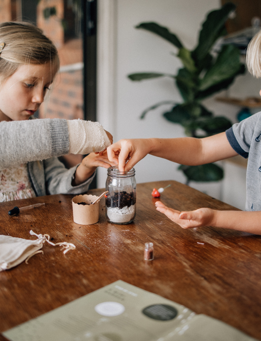 A hand is pouring soil into a Mini Garden by Poppy & Daisy. The glass jar terrarium contains layers of sand, rocks, soil, and small green plants, with a decorative mushroom and ladybug inside. Other similar Mini Gardens can be seen in the background.