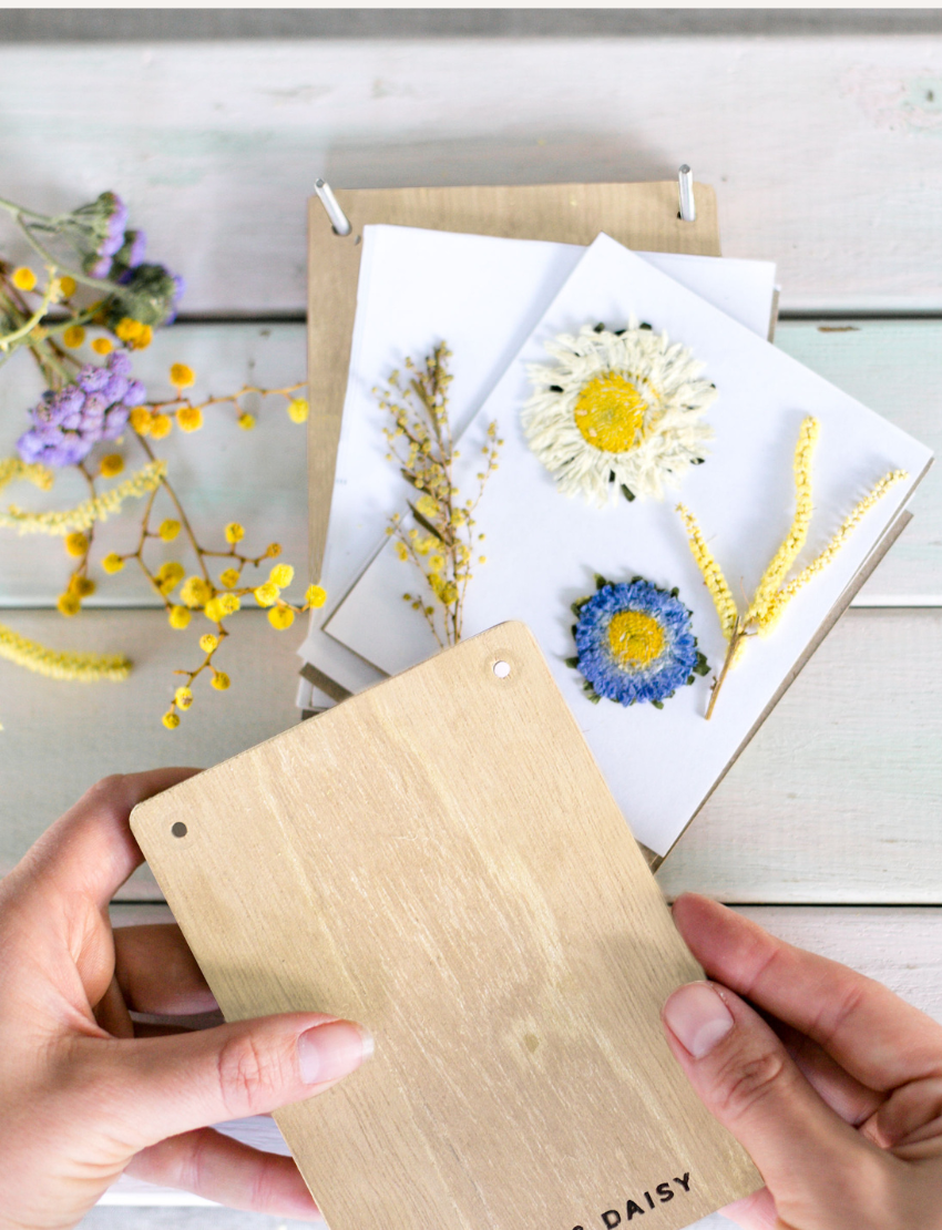 The Poppy & Daisy Flower Press kit is perfect for creating pressed flowers and includes wooden boards engraved with "Poppy & Daisy" text, a small jar, screws, a pencil, a blank round keychain as a unique keepsake, and a craft envelope. The packaging features an illustrated label with a floral pressing design.