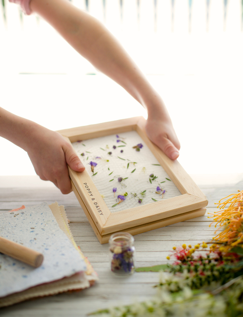 Hands gently cradle a wooden frame embellished with flowers and leaves, showcasing the process of creating Poppy & Daisy's Handmade Paper. A small jar brimming with flowers sits alongside an array of vibrant sheets of this colorful handmade paper on the table, all bathed in bright natural light.