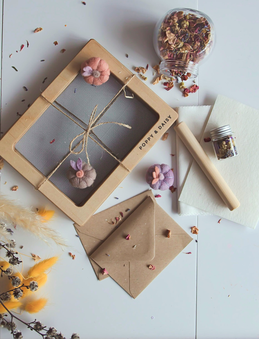 Hands gently cradle a wooden frame embellished with flowers and leaves, showcasing the process of creating Poppy & Daisy's Handmade Paper. A small jar brimming with flowers sits alongside an array of vibrant sheets of this colorful handmade paper on the table, all bathed in bright natural light.