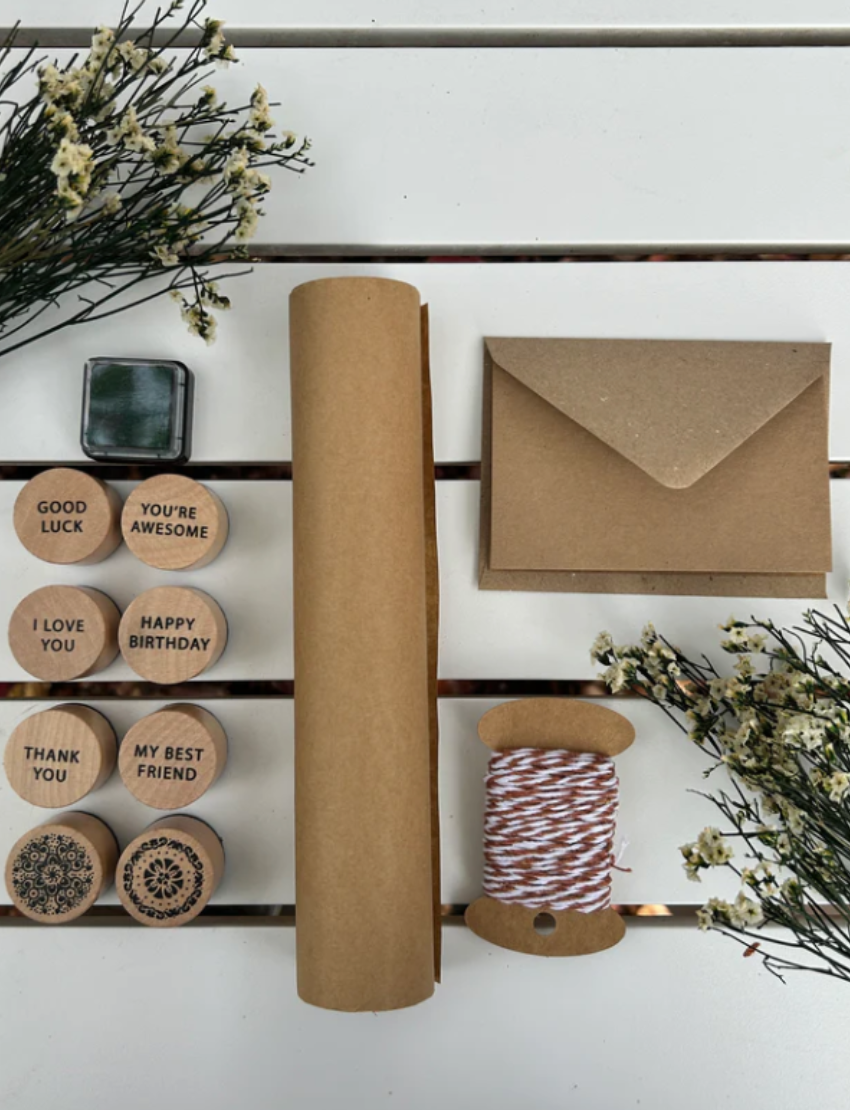 A flat lay display featuring the Poppy & Daisy DIY Gifting Set on a white surface showcases an enchanting collection for a delightful crafting experience. This set includes four rubber stamps with various messages, a vibrant green ink pad, a roll of brown paper, handmade cards with matching envelopes, a spool of twine, and small branches adorned with delicate white flowers.