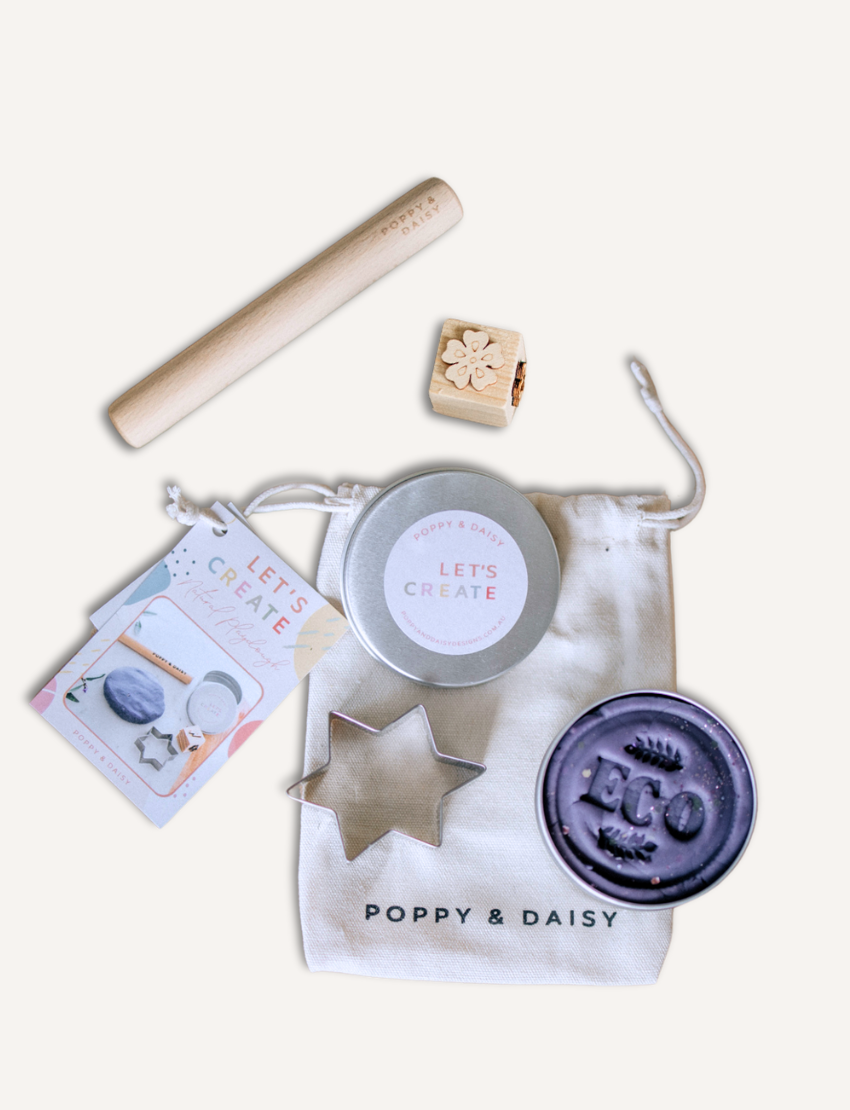 A child in a striped shirt is playing with gray Natural Playdough from Poppy & Daisy. On the table, there is a wooden stamp featuring star and flower designs and a closed metal tin labeled "Let's Create." The background is plain and light-colored.