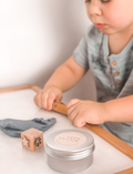 A child in a striped shirt is playing with gray Natural Playdough from Poppy & Daisy. On the table, there is a wooden stamp featuring star and flower designs and a closed metal tin labeled 