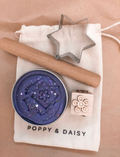 A child in a striped shirt is playing with gray Natural Playdough from Poppy & Daisy. On the table, there is a wooden stamp featuring star and flower designs and a closed metal tin labeled 