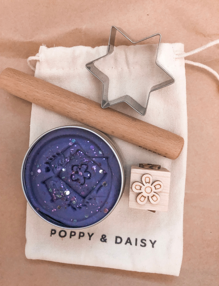 A child in a striped shirt is playing with gray Natural Playdough from Poppy & Daisy. On the table, there is a wooden stamp featuring star and flower designs and a closed metal tin labeled "Let's Create." The background is plain and light-colored.