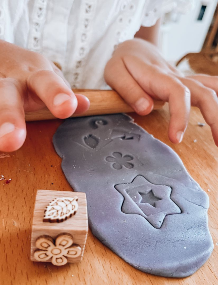 A child in a striped shirt is playing with gray Natural Playdough from Poppy & Daisy. On the table, there is a wooden stamp featuring star and flower designs and a closed metal tin labeled "Let's Create." The background is plain and light-colored.