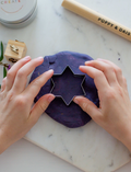 A child in a striped shirt is playing with gray Natural Playdough from Poppy & Daisy. On the table, there is a wooden stamp featuring star and flower designs and a closed metal tin labeled 