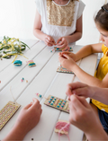 Children are seated at a white table, engaged in crafting activities using the Poppy & Daisy DIY Bookmark kit. They are threading colorful beads onto strings attached to cardboards. One child is wearing a white shirt with a gold sequin front, while another sports a yellow shirt. The table is adorned with flowers.