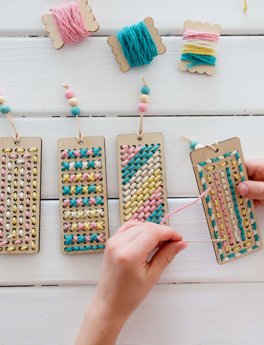 Children are seated at a white table, engaged in crafting activities using the Poppy & Daisy DIY Bookmark kit. They are threading colorful beads onto strings attached to cardboards. One child is wearing a white shirt with a gold sequin front, while another sports a yellow shirt. The table is adorned with flowers.