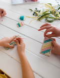 Children are seated at a white table, engaged in crafting activities using the Poppy & Daisy DIY Bookmark kit. They are threading colorful beads onto strings attached to cardboards. One child is wearing a white shirt with a gold sequin front, while another sports a yellow shirt. The table is adorned with flowers.