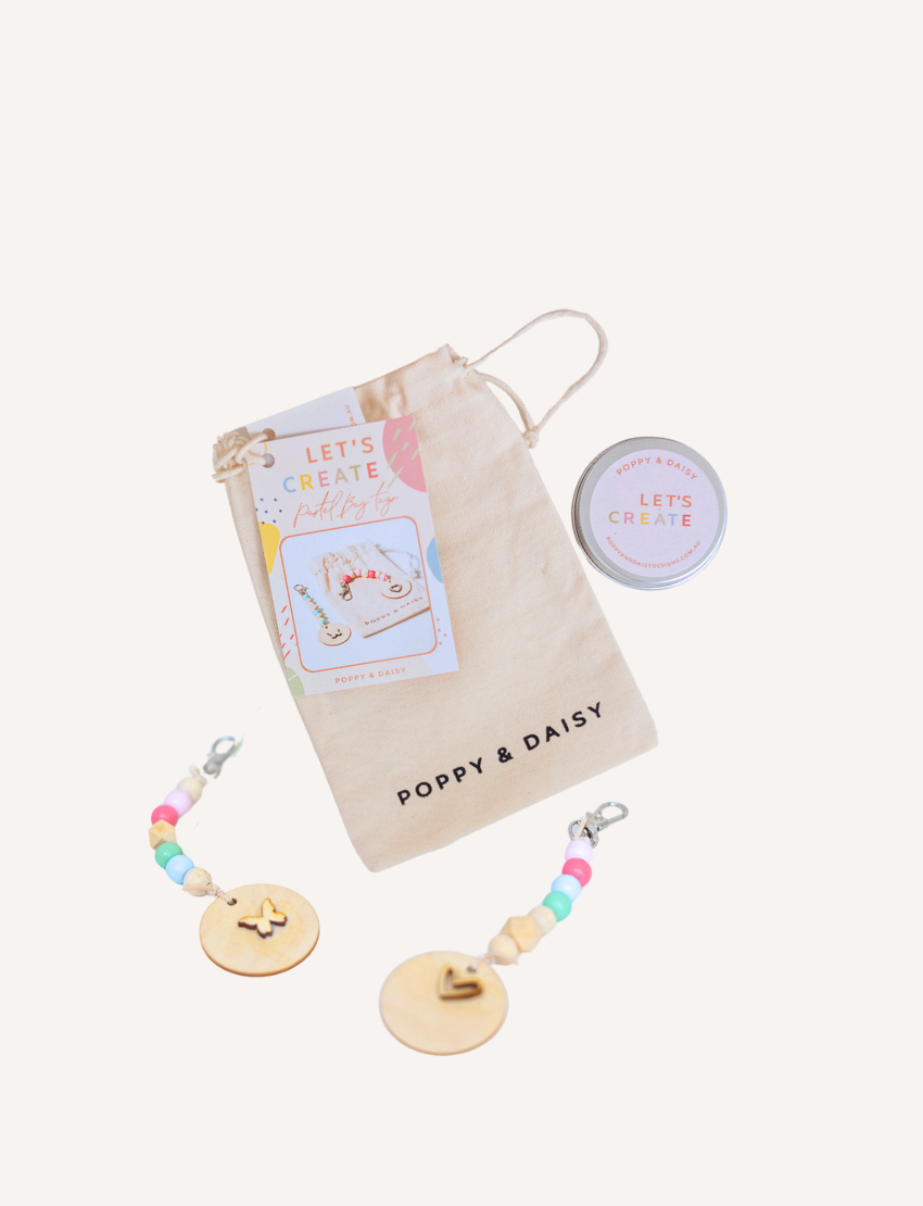 A person is decorating a Pastel Bag Tag from Poppy & Daisy, painting a heart shape with a small brush on the wooden disc. The tag rests on a wooden surface, and to the right, an open tin filled with colorful beads can be seen. A string and part of a pouch appear on the left.