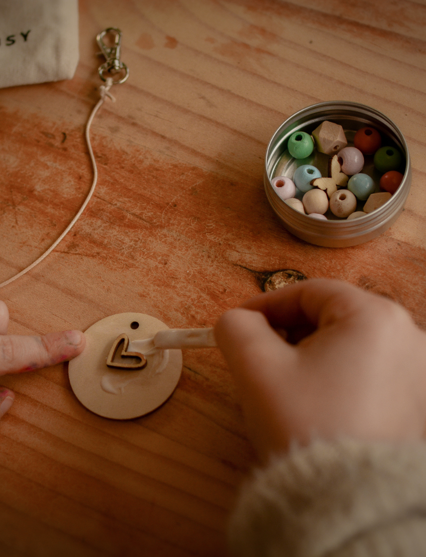 A person is decorating a Pastel Bag Tag from Poppy & Daisy, painting a heart shape with a small brush on the wooden disc. The tag rests on a wooden surface, and to the right, an open tin filled with colorful beads can be seen. A string and part of a pouch appear on the left.