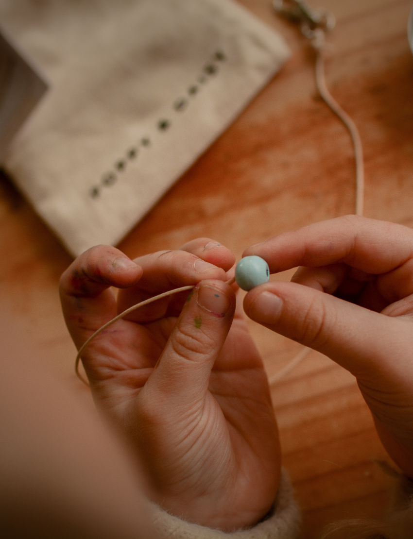 A person is decorating a Pastel Bag Tag from Poppy & Daisy, painting a heart shape with a small brush on the wooden disc. The tag rests on a wooden surface, and to the right, an open tin filled with colorful beads can be seen. A string and part of a pouch appear on the left.