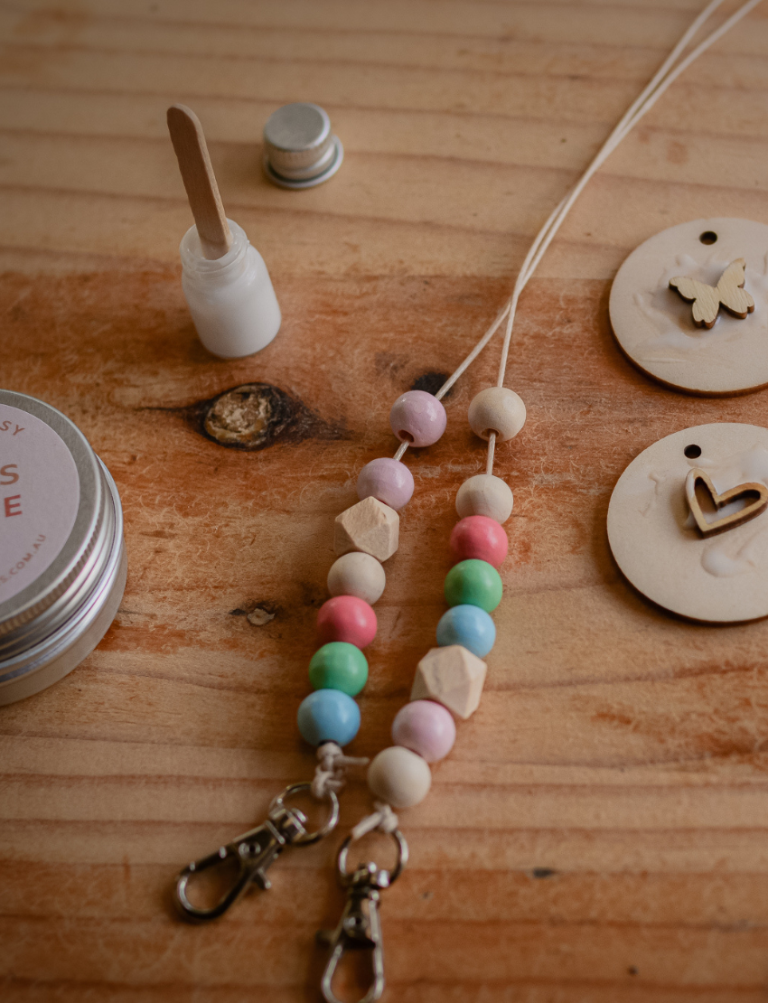 A person is decorating a Pastel Bag Tag from Poppy & Daisy, painting a heart shape with a small brush on the wooden disc. The tag rests on a wooden surface, and to the right, an open tin filled with colorful beads can be seen. A string and part of a pouch appear on the left.