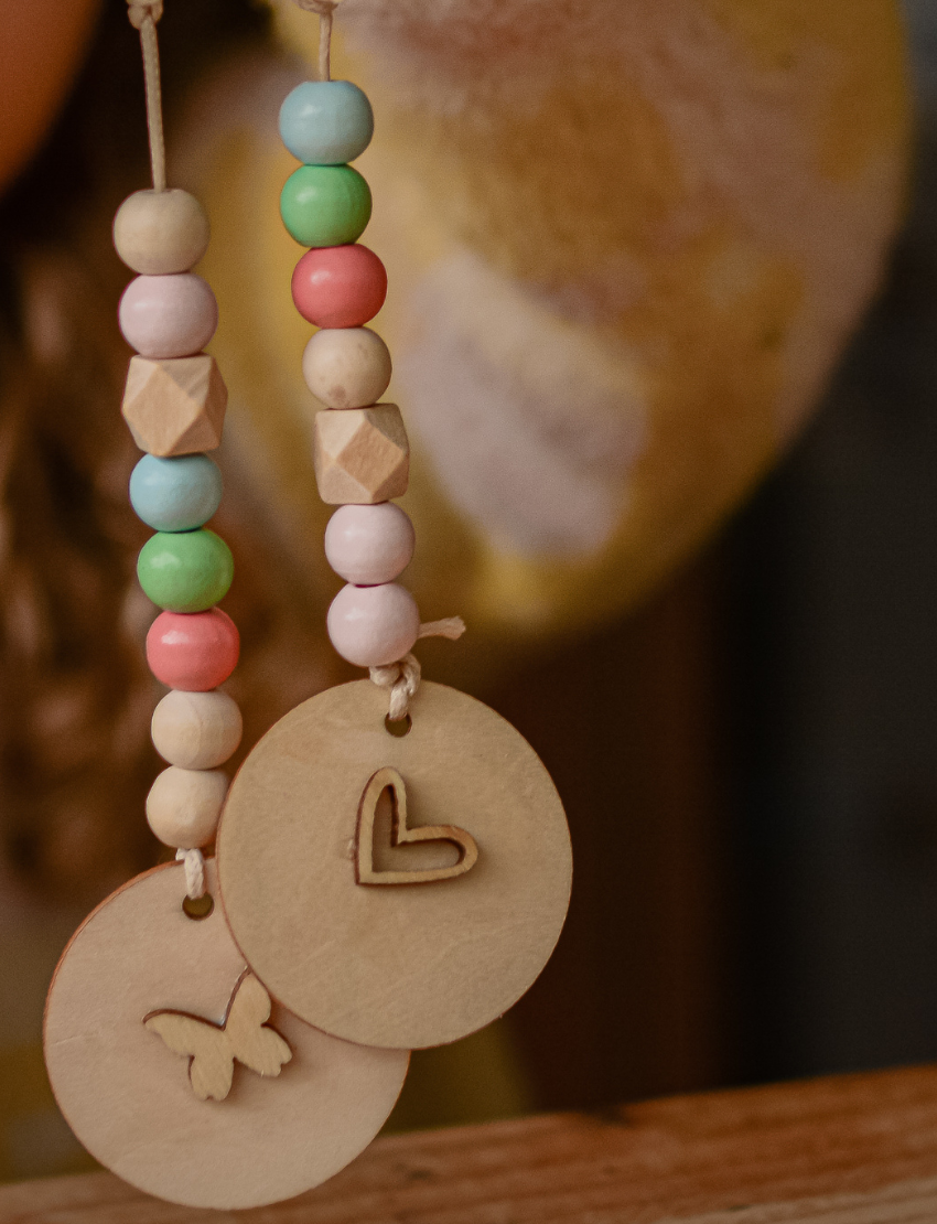A person is decorating a Pastel Bag Tag from Poppy & Daisy, painting a heart shape with a small brush on the wooden disc. The tag rests on a wooden surface, and to the right, an open tin filled with colorful beads can be seen. A string and part of a pouch appear on the left.