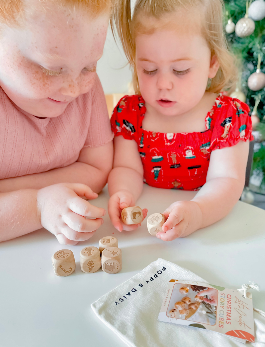 A person wearing a white sweater holds five wooden Christmas Story Cubes by Poppy & Daisy, each displaying different nature-themed images, above a round woven tray. The tray is filled with pinecones and red flowers, set on a sandy surface.