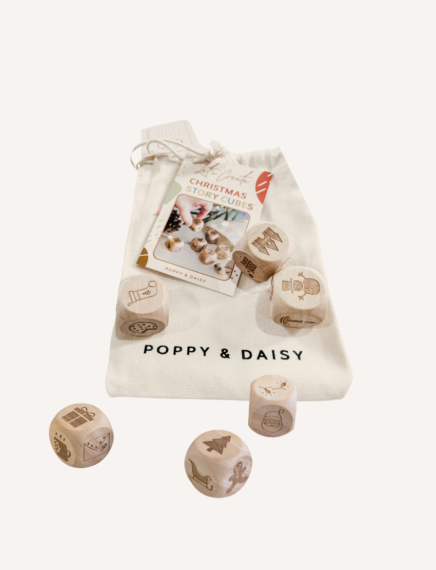 A person wearing a white sweater holds five wooden Christmas Story Cubes by Poppy & Daisy, each displaying different nature-themed images, above a round woven tray. The tray is filled with pinecones and red flowers, set on a sandy surface.