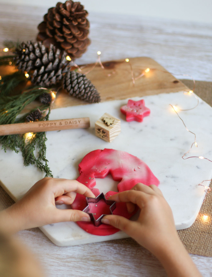 A child uses a star-shaped cutter to cut out star shapes from Poppy & Daisy's Candy Cane Playdough on a marble board, while pinecones, greenery, a wooden roller, and string lights in the background enhance the festive atmosphere.