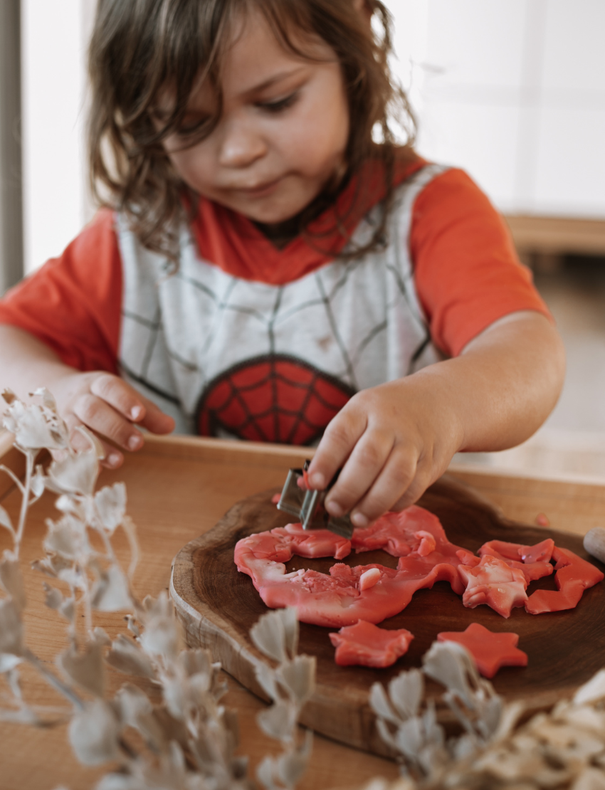 A child uses a star-shaped cutter to cut out star shapes from Poppy & Daisy's Candy Cane Playdough on a marble board, while pinecones, greenery, a wooden roller, and string lights in the background enhance the festive atmosphere.