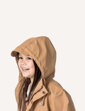 The Cry Wolf Play Jacket in tan is displayed against a white background. The raincoat features a front button closure, drawstrings on the hood, two front pockets with button flaps, and long sleeves with elastic cuffs. A small logo is visible on the left chest area.