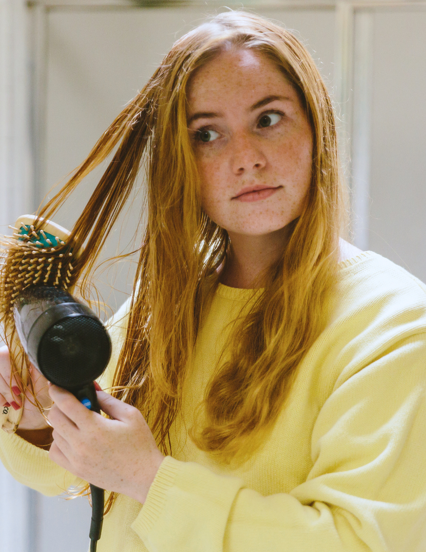 A person with long red hair wearing a yellow sweater blow-dries their hair using a round brush while taking part in The Play Way's Puberty Self-Care Masterclass. They are looking slightly to the right with a focused expression within an indoor space that features a blurred wall and partial window in the background.